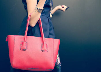 Fashionable beautiful big red handbag on the arm of the girl in a fashionable black dress, posing near the wall on a warm summer night. Warm color